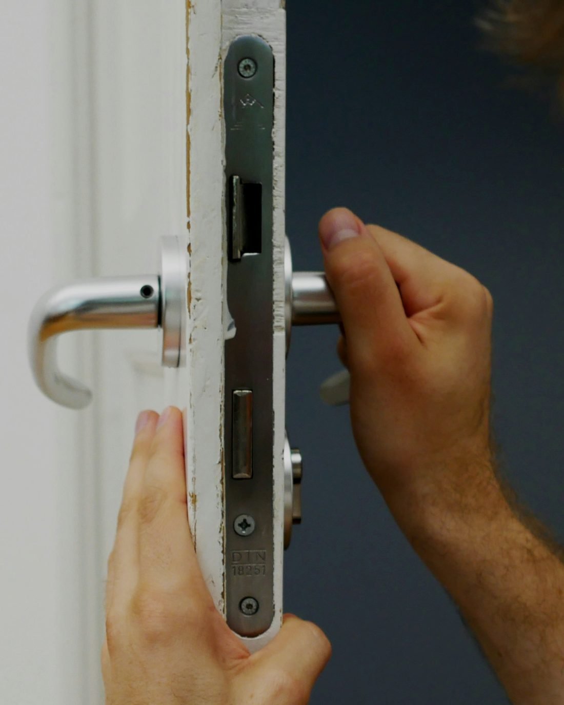 Deux mains posant une serrure à mortaise et une poignée à béquille dans le chant d’une porte blanche
