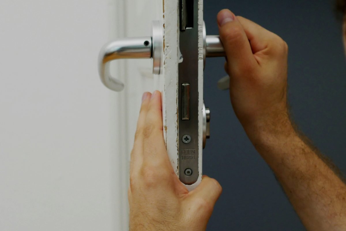 Hands adjusting a mortise lock and lever handle on the edge of a white interior door
